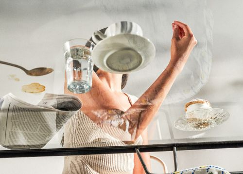 Woman's arm reaches up beneath a clear table with coffee cups, glasses, and newspaper, creating a layered, abstract composition against a bright background.