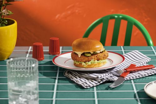 Crispy chicken sandwich served with pickles and shredded lettuce on a checkered table with red silverware and a bright orange background.