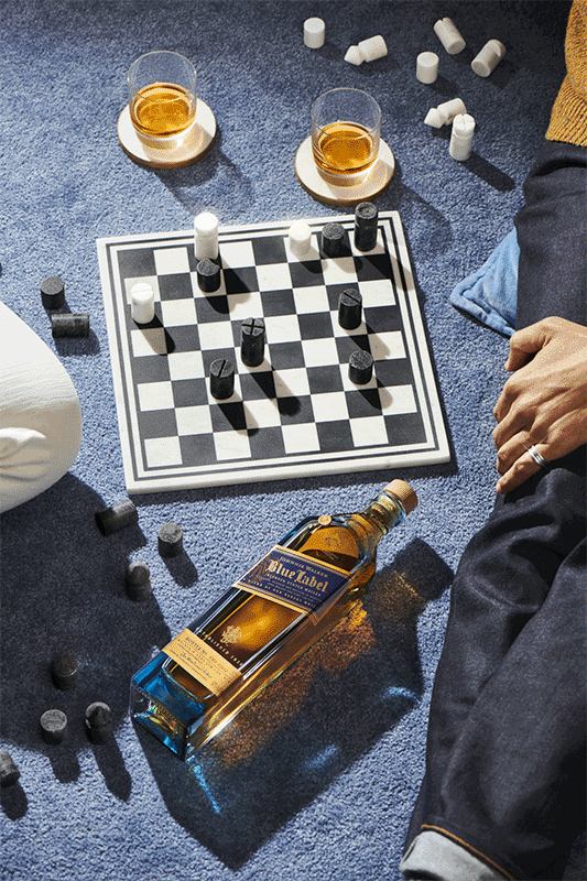 Overhead shot of a bottle of Johnnie Walker Blue Label, a chessboard, and two glasses of whiskey on a blue textured surface.