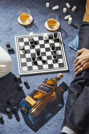 Overhead shot of a bottle of Johnnie Walker Blue Label, a chessboard, and two glasses of whiskey on a blue textured surface.