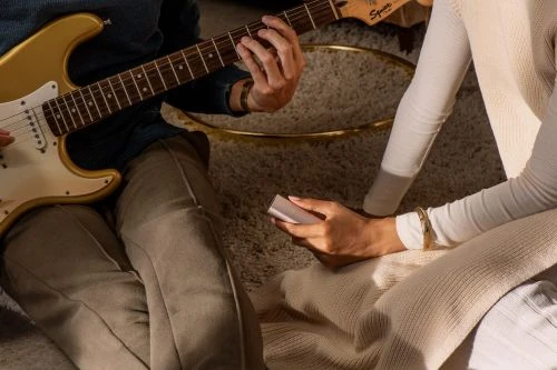 Man playing a gold electric guitar while a woman holds a smartphone, in a cozy, softly lit interior setting with a shag carpet.