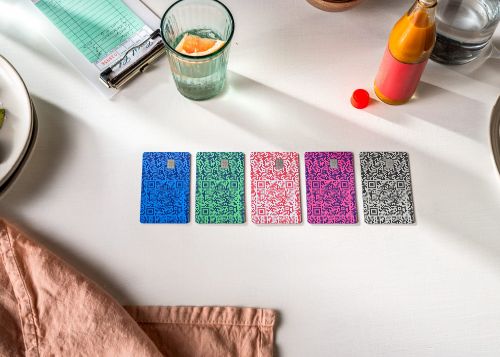 Overhead shot of six colorful QR code cards lined up on a white table, along with a drink, juice, receipt, and cloth.