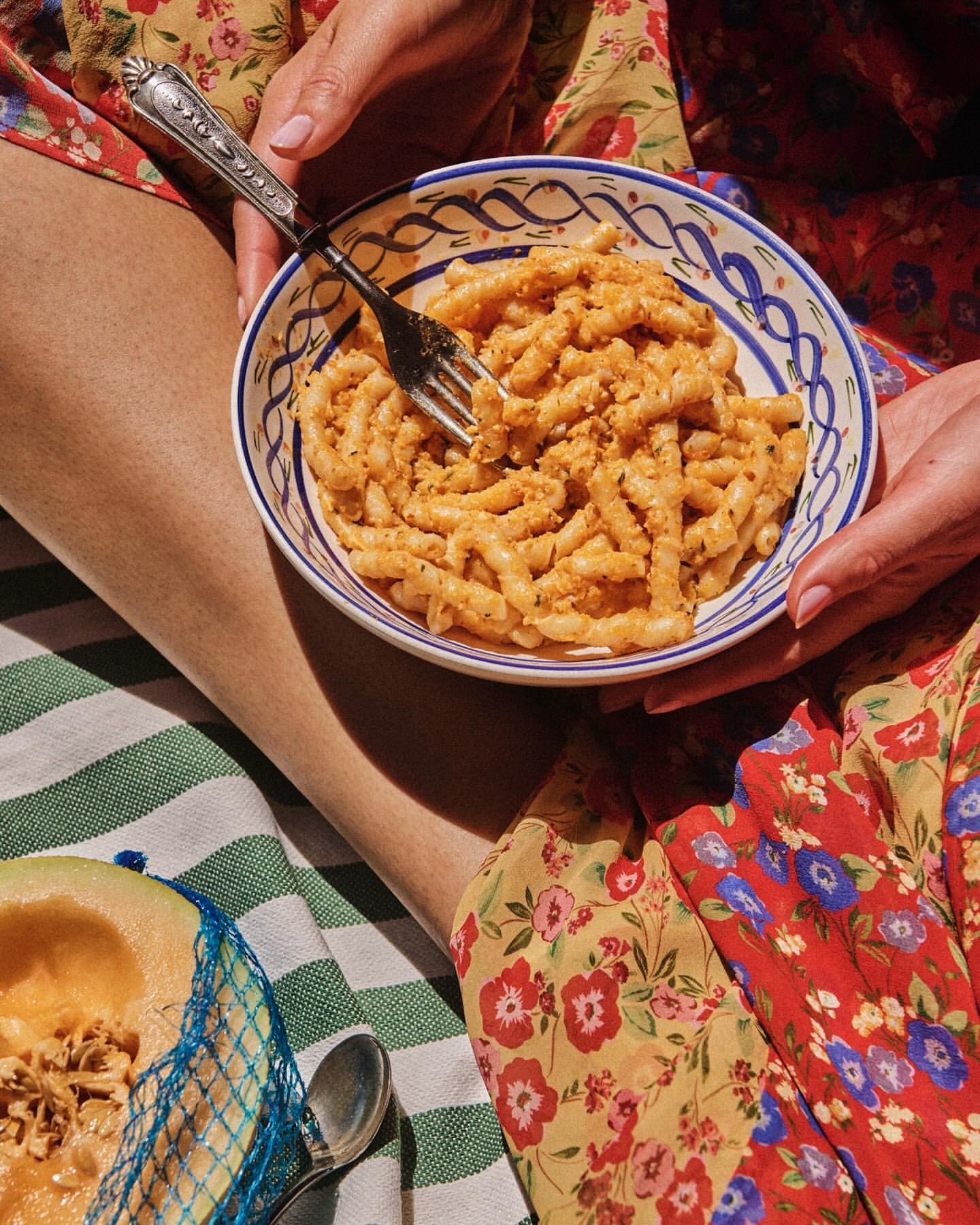 Bowl of pasta held by a person wearing a floral dress, featuring a fork, striped blanket, and melon slice in the background. 
