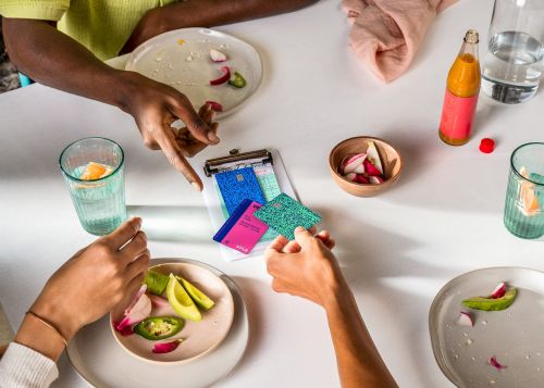 Overhead shot of two people at a white table with drinks and plates of food, with colorful cards on a clipboard.
