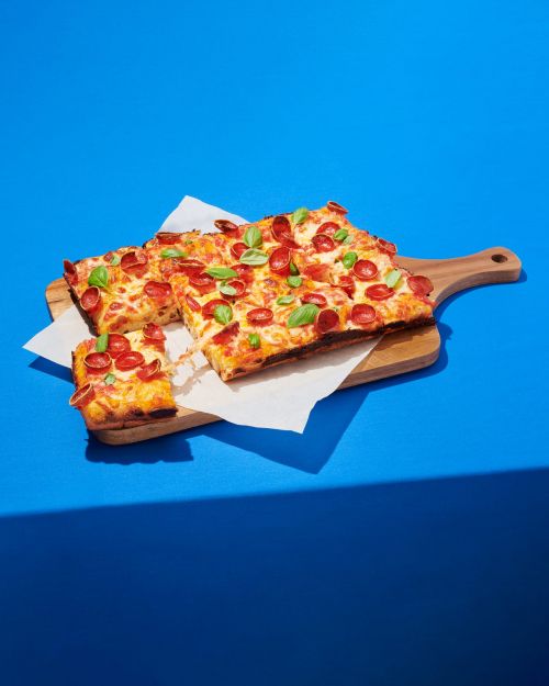 Overhead shot of square pepperoni pizzas with basil leaves on wooden cutting board, resting on white parchment against blue background.
