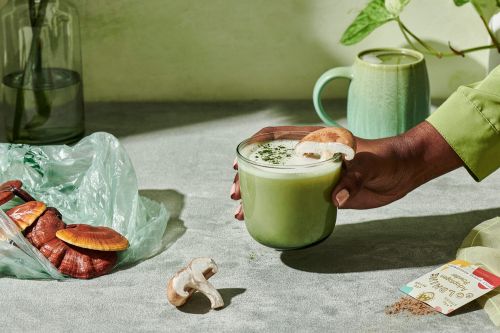 Hand holding a green drink with mushroom garnish, mushrooms in a plastic bag, green mug, and packet on a textured surface.
