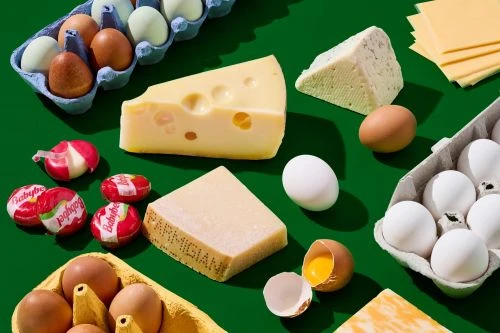 Overhead shot showcases an assortment of dairy and egg products including cheese wedges, Babybel cheeses, and cartons of chicken eggs on green.