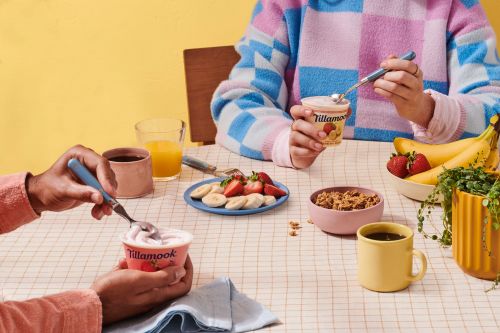 Two people at a breakfast table with yogurt, fruit, granola, juice, and coffee, showcasing a brightly lit and colourful breakfast scene.