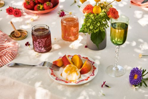 A still life with jars of jam, peaches, strawberries, and flowers, set on a white tablecloth with soft, natural lighting.