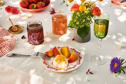 A still life with jars of jam, peaches, strawberries, and flowers, set on a white tablecloth with soft, natural lighting.
