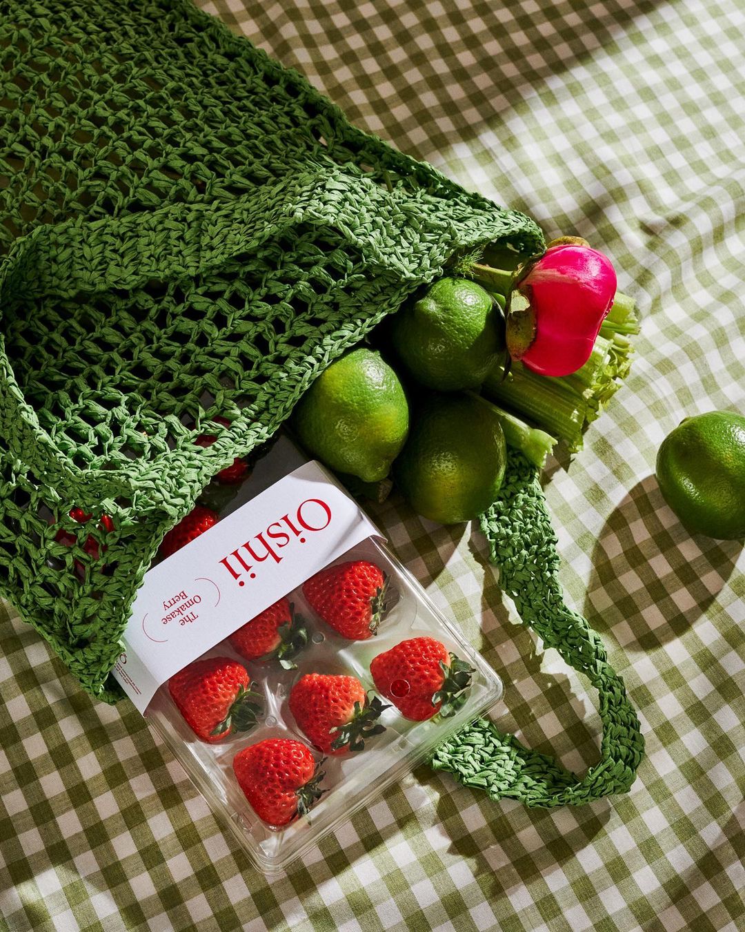 Close-up of a green woven bag filled with strawberries in a container, limes, a radish, and celery on a green and white checkered tablecloth.