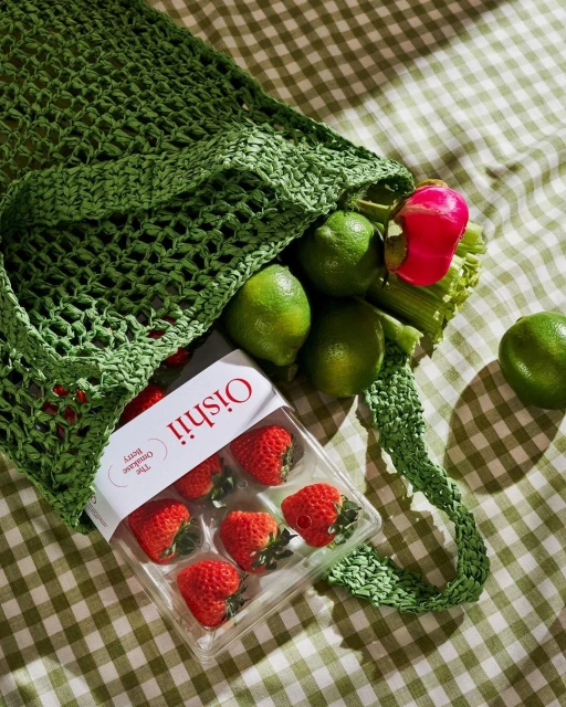 Close-up of a green woven bag filled with strawberries in a container, limes, a radish, and celery on a green and white checkered tablecloth.