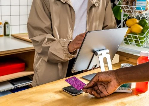 A customer uses a contactless card to pay at a cafe. The cashier holds a digital tablet in an indoor setting with natural light.