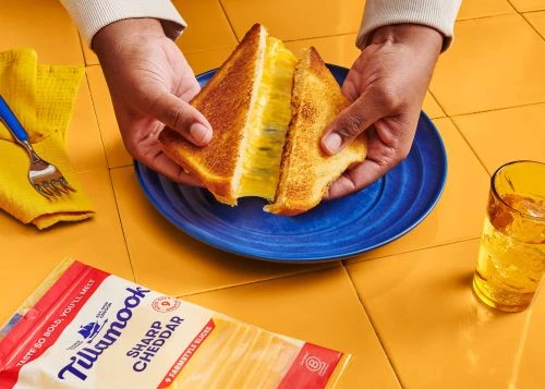 Grilled cheese sandwich being held over blue plate on yellow tile with Tillamook cheese package and a glass of iced beverage.