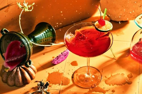 Artistic studio shot of a red cocktail in a stemmed glass, garnished with a cherry and lime, beside exotic fruit and decorative vase.