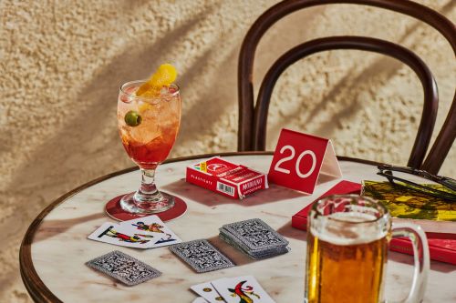 A cocktail with a garnish and a beer sit on a marble table with playing cards scattered around, creating a relaxed, social atmosphere.