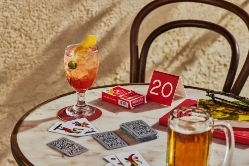A cocktail with a garnish and a beer sit on a marble table with playing cards scattered around, creating a relaxed, social atmosphere.
