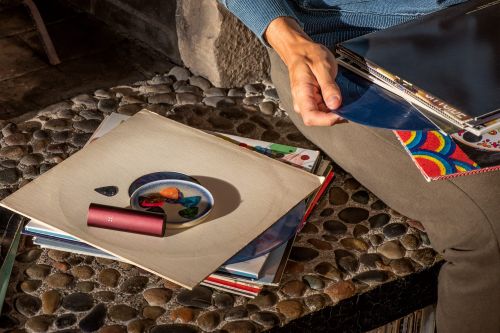 Close-up of a person sitting by a stone wall, holding vinyl records, with a pipe and other objects on a record sleeve.