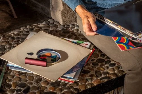 Close-up of a person sitting by a stone wall, holding vinyl records, with a pipe and other objects on a record sleeve.