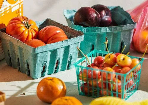 Close-up of fresh fruits and vegetables in blue paper containers: tomatoes, plums, cherries, and oranges on a white surface with natural lighting.