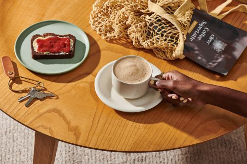 Overhead shot of an unidentifiable person holding a cup of coffee, beside a piece of jam-covered toast and keys, on a wooden table.