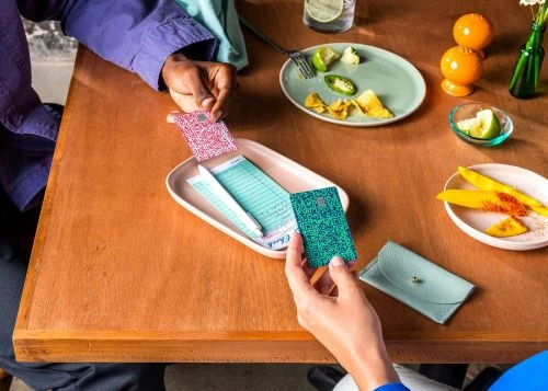 Two people at a wooden table with a check tray, credit cards, a coin purse, plates of fruit, chips, lime, and a drink.