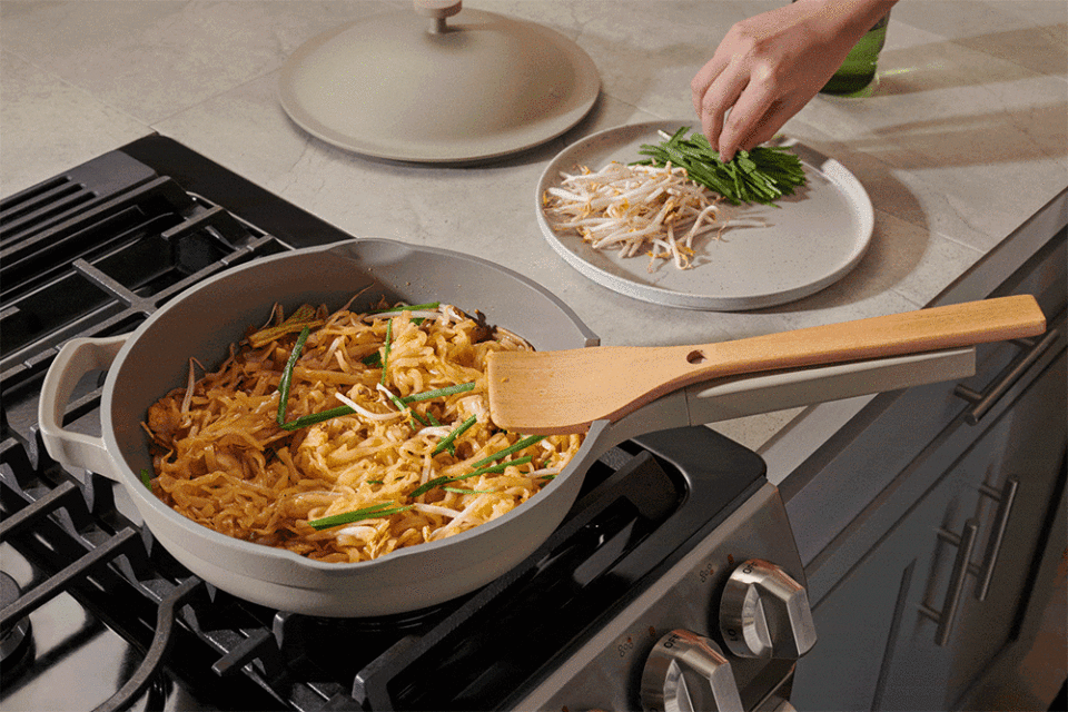Pad Thai being prepared on a stove top with a wooden spatula, garnished with green onions and bean sprouts on a plate.