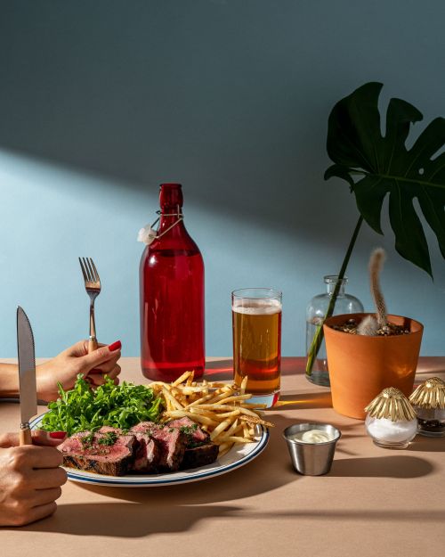 Overhead shot of a sliced steak on a plate with fries and greens, alongside a red bottle, beer, and a potted plant on a table.