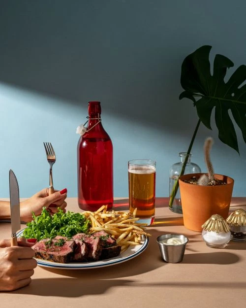 Overhead shot of a sliced steak on a plate with fries and greens, alongside a red bottle, beer, and a potted plant on a table.