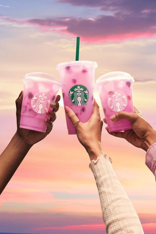 Three individuals raise Starbucks beverages with pink liquid towards a vibrant sunset sky, highlighting a celebratory, refreshing moment.