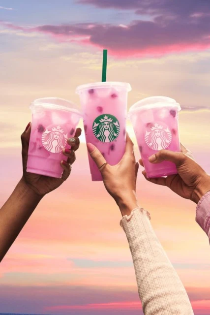 Three individuals raise Starbucks beverages with pink liquid towards a vibrant sunset sky, highlighting a celebratory, refreshing moment.
