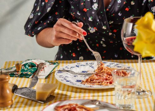 Woman enjoys spaghetti with tomato sauce at a table set with wine, cheese, silverware, and a checkered tablecloth, indoor food setting.