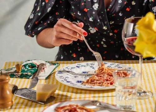 Woman enjoys spaghetti with tomato sauce at a table set with wine, cheese, silverware, and a checkered tablecloth, indoor food setting.