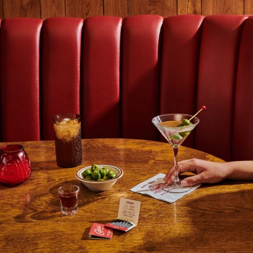 Martini and soft drink on a wooden table with olives, a matchbook, and a patterned coaster in a dimly lit restaurant.