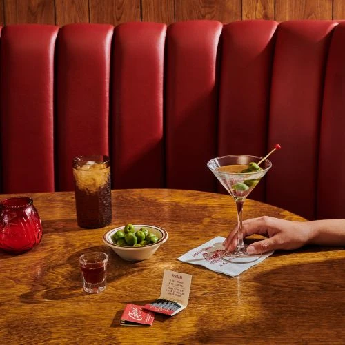 Martini and soft drink on a wooden table with olives, a matchbook, and a patterned coaster in a dimly lit restaurant.
