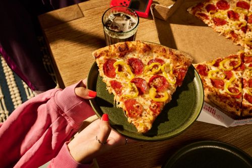 A slice of pepperoni pizza on a plate held by hands with red nail polish, with the pizza box and a drink visible.