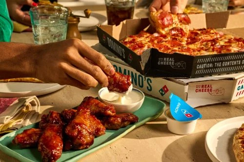 Overhead shot of Domino's pizza and chicken wings, featuring a hand dipping a wing in sauce, soda glasses, and Ranch sauce.
