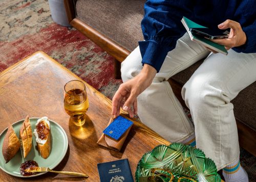 Overhead shot of a person seated at a wooden table with a drink, bread, jam, wallet, credit card, and passport displayed.