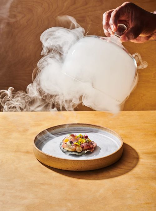 A dish of food is presented on a plate with vapor or smoke being held above it by a hand against a warm brown background.