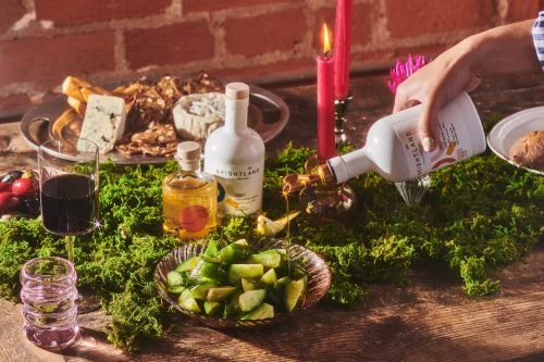 Olive oil being poured onto a salad with cheese and crackers, complemented by candles on a moss-covered wooden table.