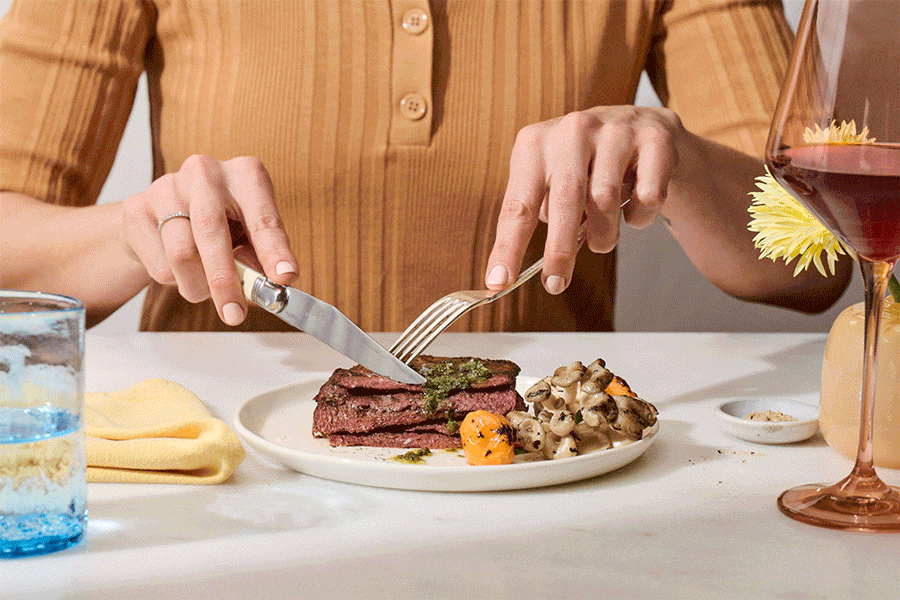 Overhead view of a person using a knife and fork to cut into a steak served with vegetables on a plate in an indoor setting.