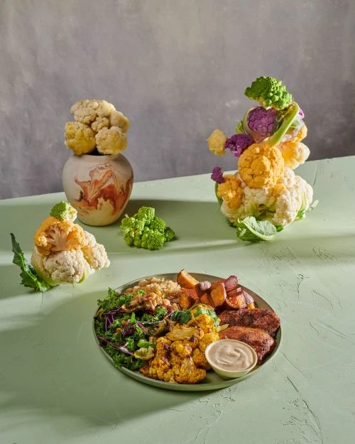 Colourful vegetarian buddha bowl with roasted vegetables and salad, with various cauliflower heads in the background, studio shot.