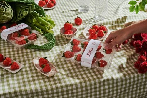 Still life with strawberries in containers and on plates, arranged on a green and white checkered tablecloth with greenery in the background.