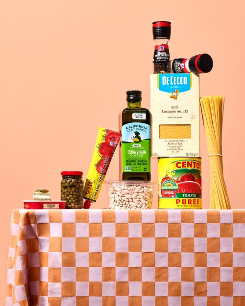Assortment of dry pasta and Italian cooking ingredients stacked on a checkered tablecloth against a peach-colored background.
