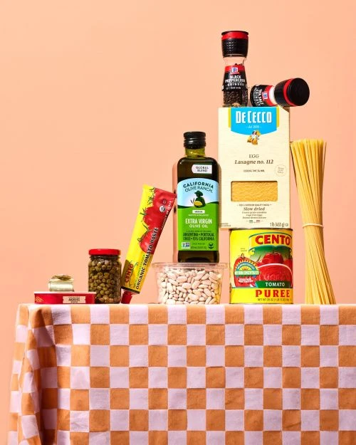 Assortment of dry pasta and Italian cooking ingredients stacked on a checkered tablecloth against a peach-colored background.