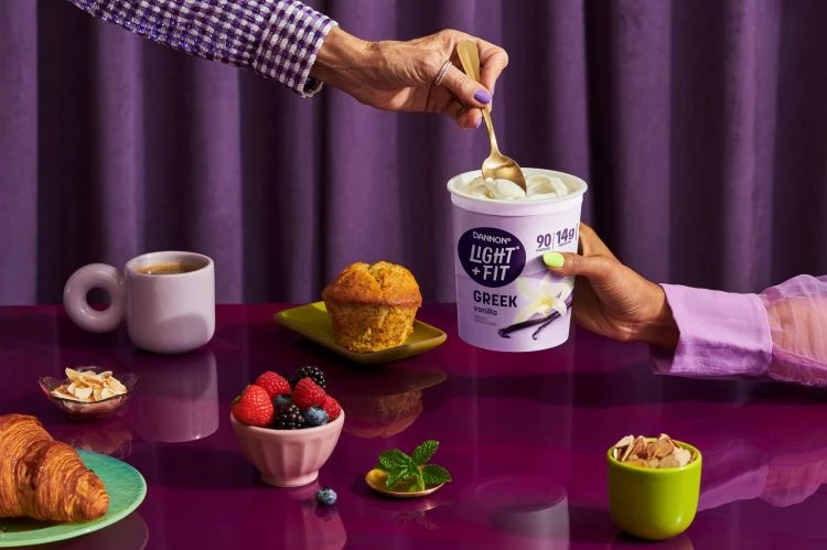 Overhead shot of a Dannon Light + Fit Greek vanilla yogurt cup being served with a gold spoon, surrounded by breakfast foods on a table.