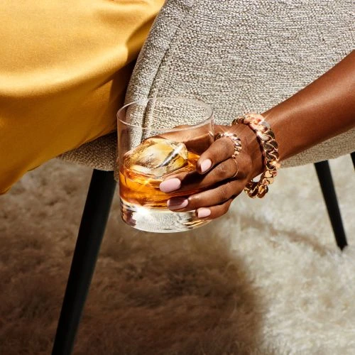 Close-up of an adult's manicured hand holding a whiskey glass filled with amber liquid and ice in an indoor setting, with soft lighting.