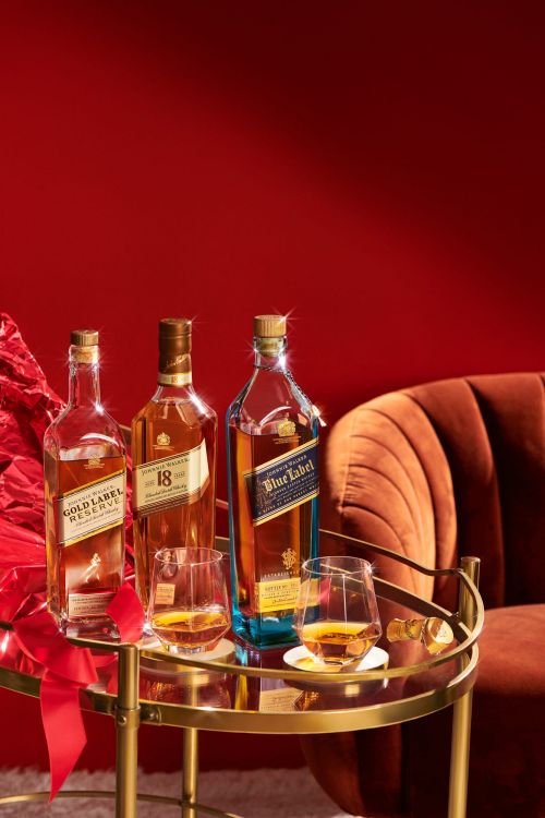 Three Johnnie Walker bottles (Gold Label, 18 Year, Blue Label) and two glasses with whiskey on a golden bar cart against red background.