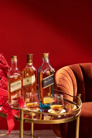 Three Johnnie Walker bottles (Gold Label, 18 Year, Blue Label) and two glasses with whiskey on a golden bar cart against red background.