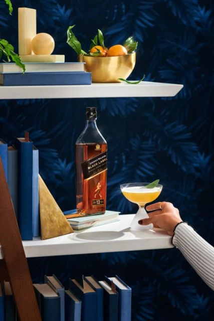 Bottle of Johnnie Walker Black Label whiskey and cocktail glass on a shelf, against a blue patterned background.